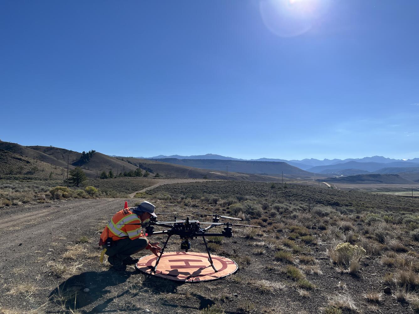 USGS drone pilot squatting by craft on landing pad located along dirt road with mountains in the distance.