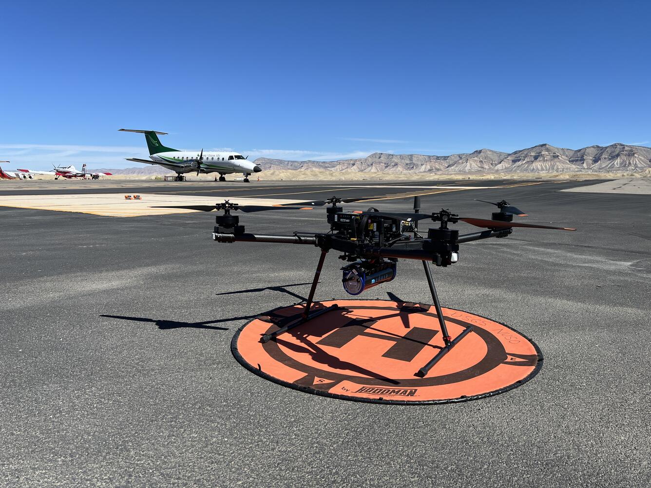 UAS sitting on landing pad at the Grand Junction Regional Airport, with small airplane in background.