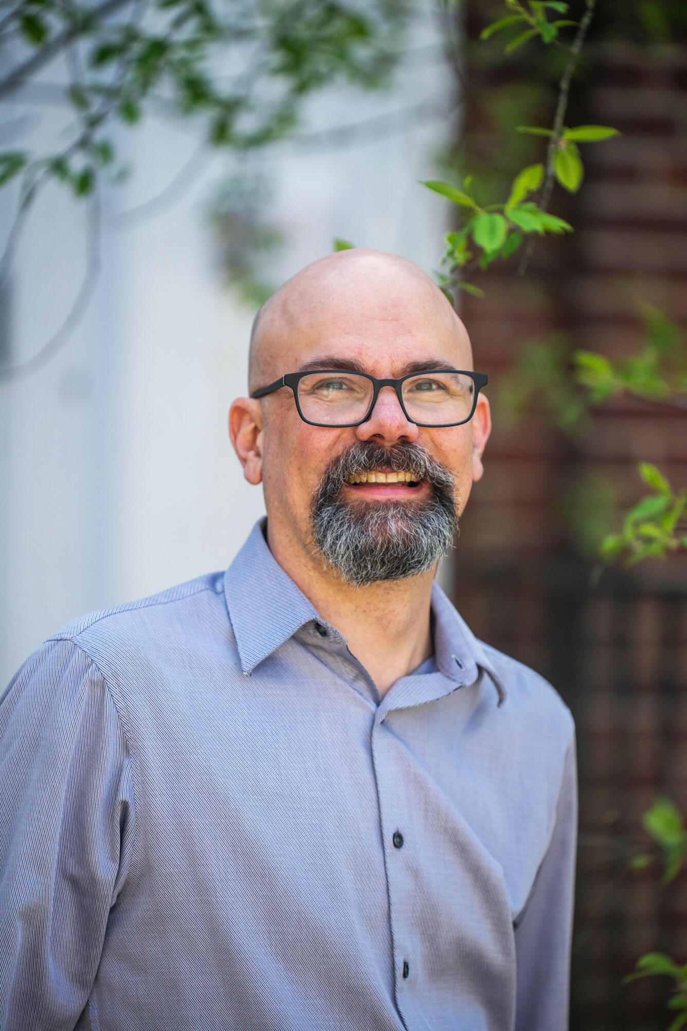 Man wearing a blue button-down shirt standing outside with tree behind him