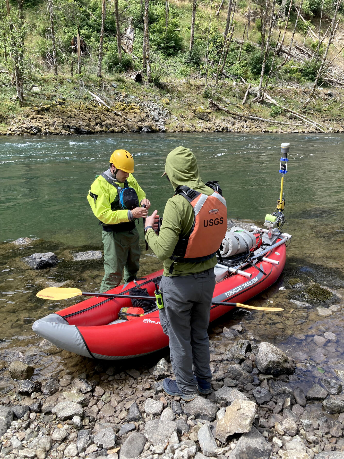 Two researchers standing near their equipment laden kayak on the rocky river bank. Green blue river water moves swiftly by