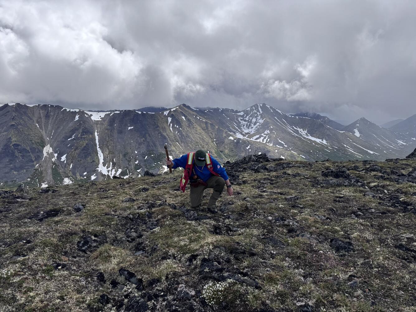 A man kneeling and holding a hammer on an alpine ridge.