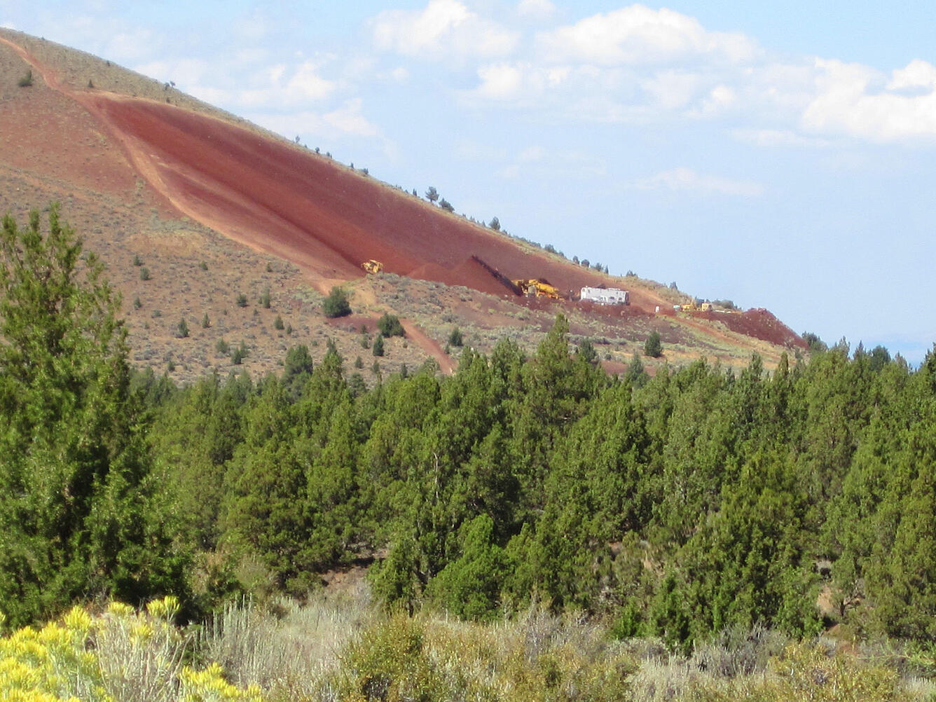 Photo of trucks mining the side of a hill, trees in the foreground.