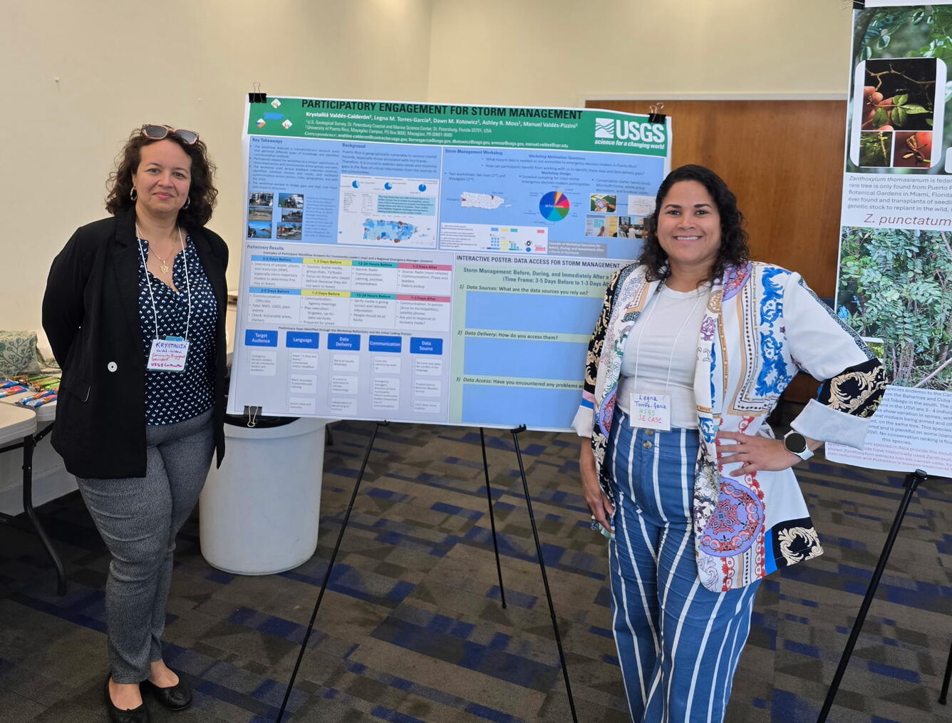 Two women stand in front of a poster on a stand.