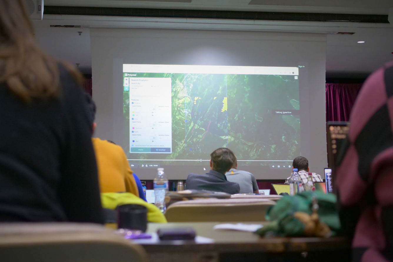 Participants of a workshop, seen from behind, look at a screen showing a large map outlining potential critical mineral data 