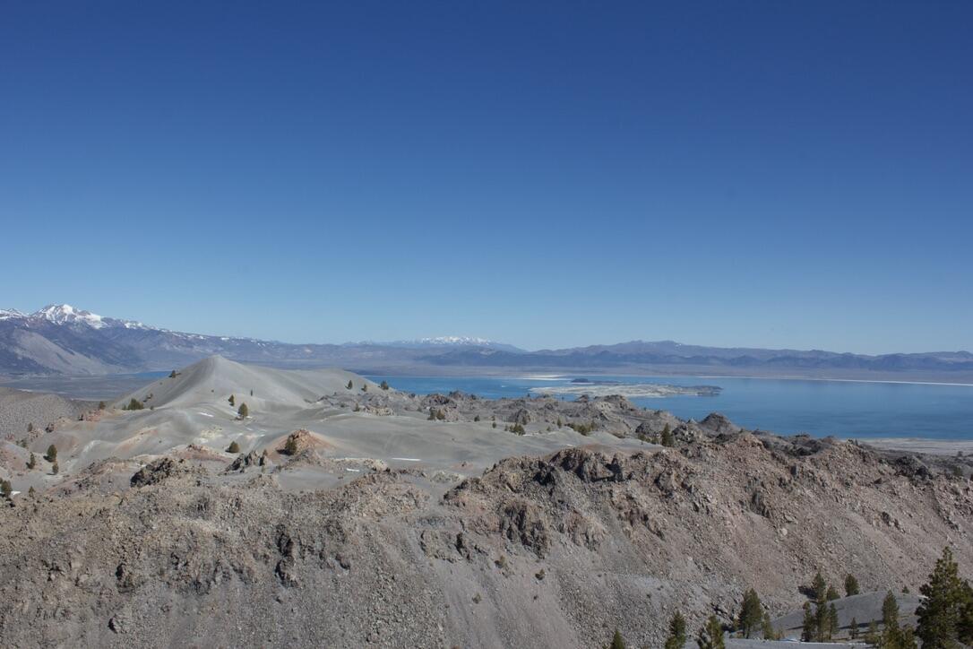 Viewed from overhead, the gray and rubbly rhyolite lava domes of the Mono-Inyo Craters are interspersed with smooth patches of volcanic ash and scoria. In the distance, a snow-capped mountain range surrounds a broad lake.