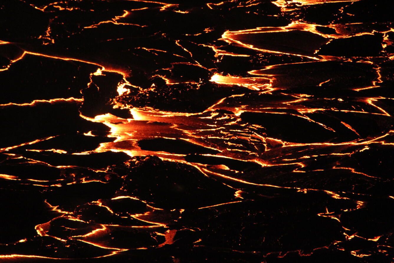 Low-light zoomed in view of lava flow with incandescent orange cracks between plates black lava crust