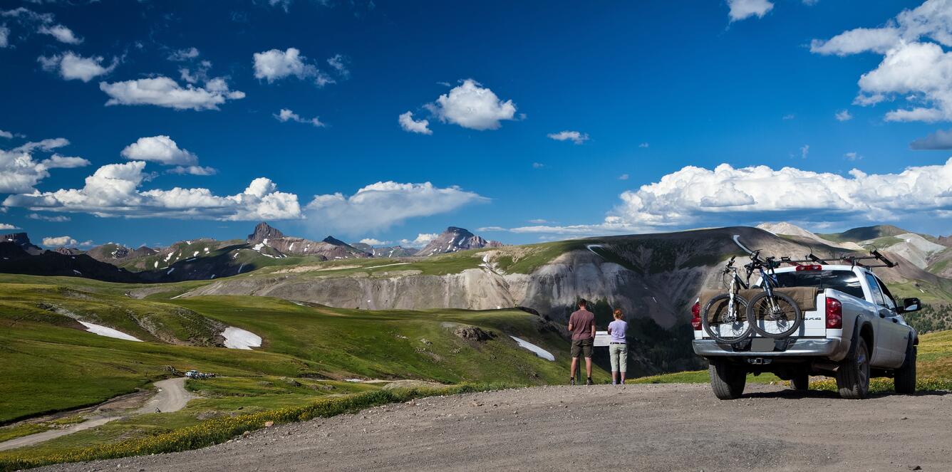 two people stand next to their car, reading a sign, with wide mountain views and partly cloudy skies in front of them