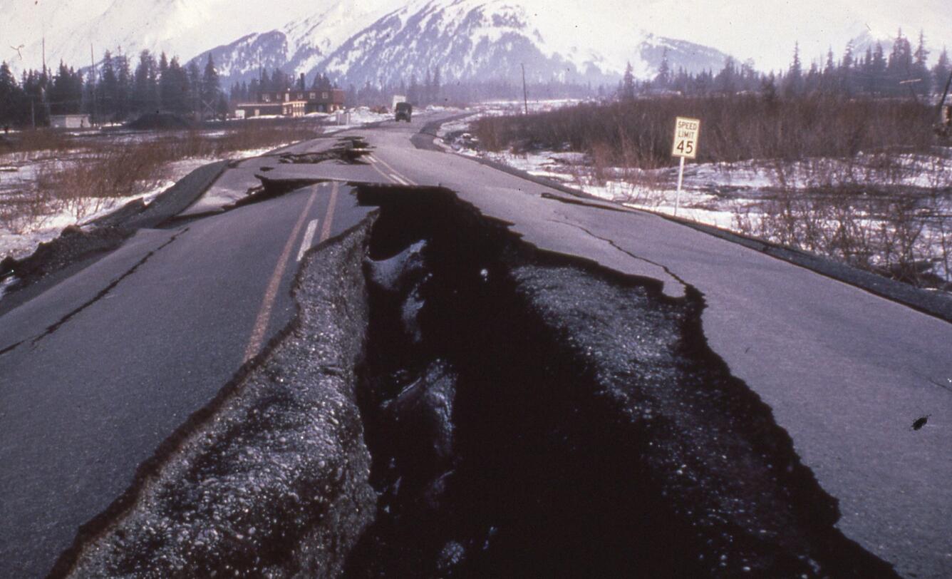 Large deep cracks opened along highway. Speed sign right side, vegetation, trees and convoy truck approaching crack.