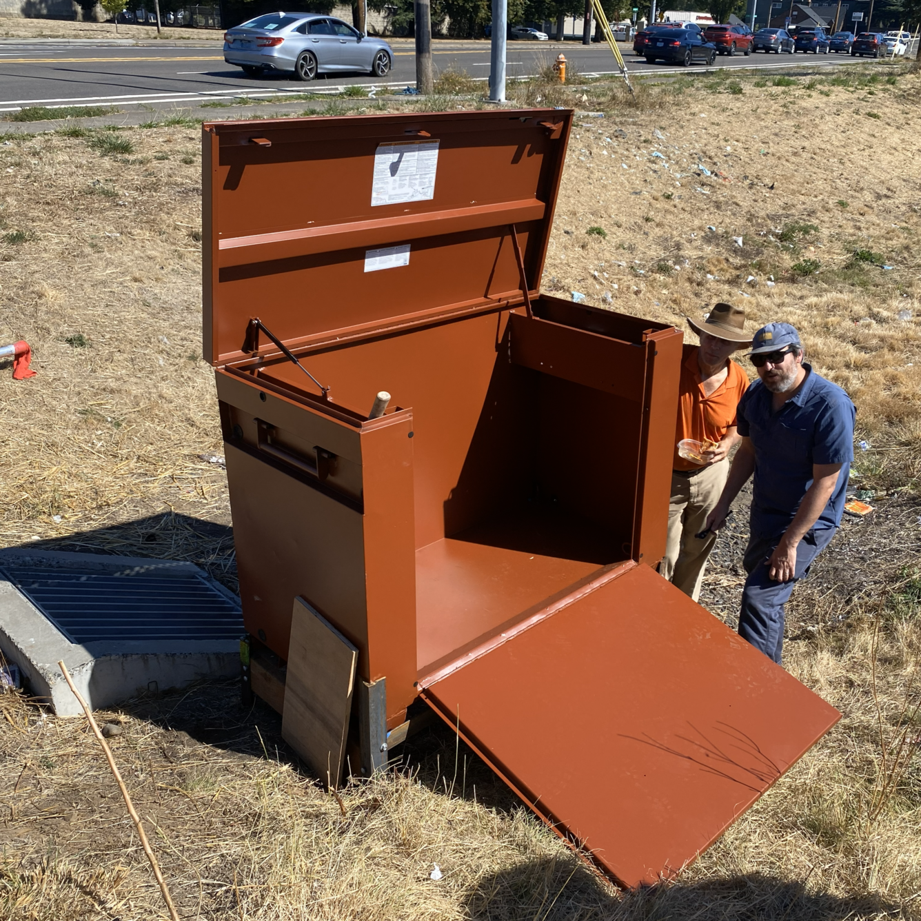two men in hat and glasses stand by a large brown metal box that is taller than them. In a ditch by a roadway.