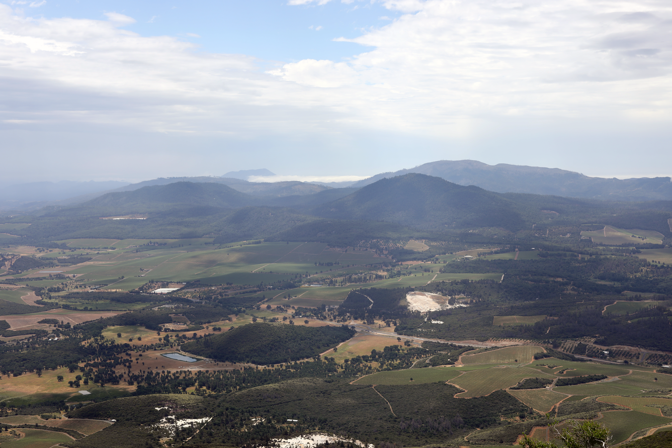 Viewed from the top of a mountain, a patchwork valley of vineyards and low rolling hills rises toward several low mountains in the distance. Several mines in the foreground reveal bright white altered volcanic rock.