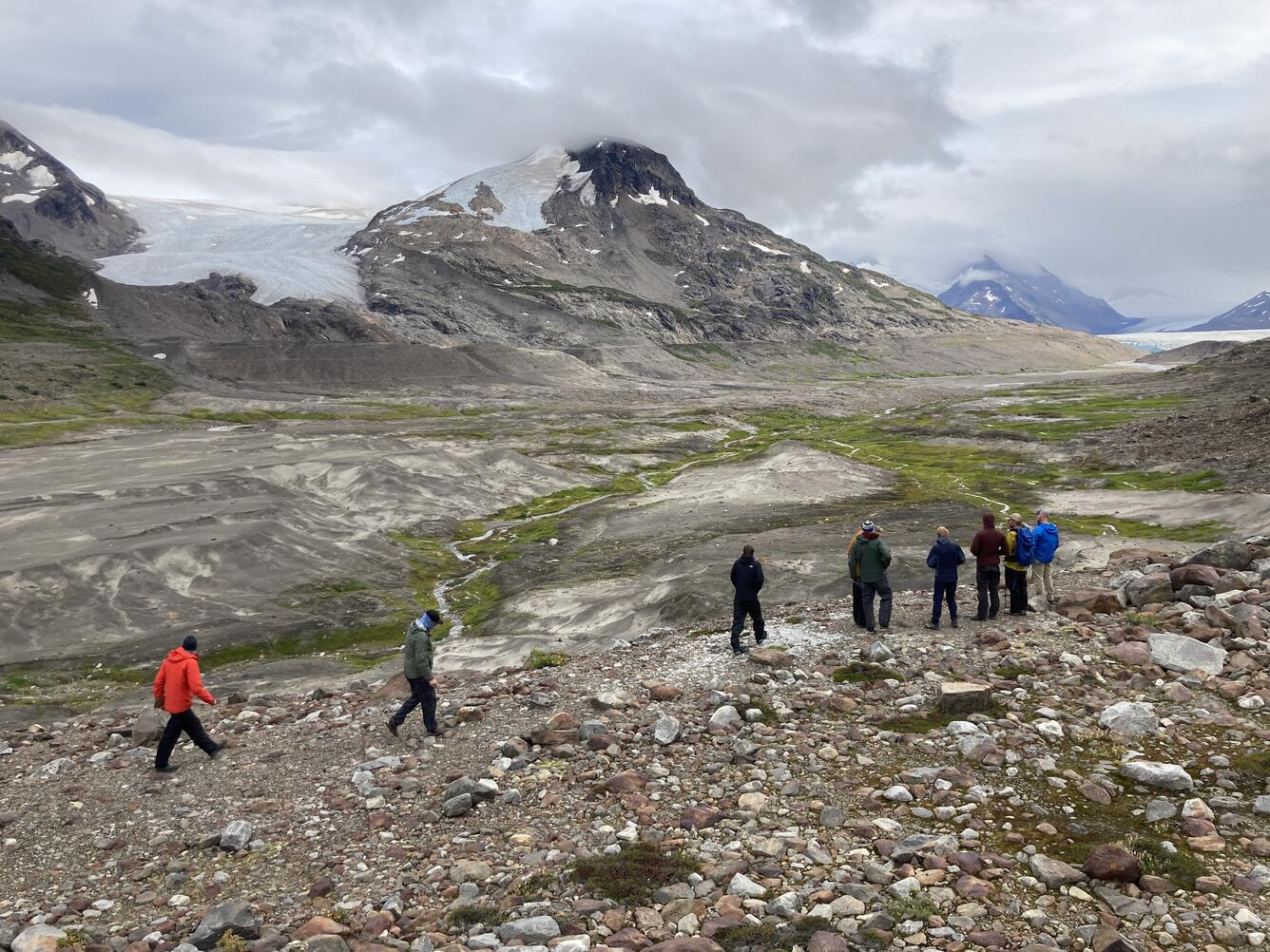 Nine scientists standing on rocky land. Stream below with ice covered mountains and iceberg lake to right.