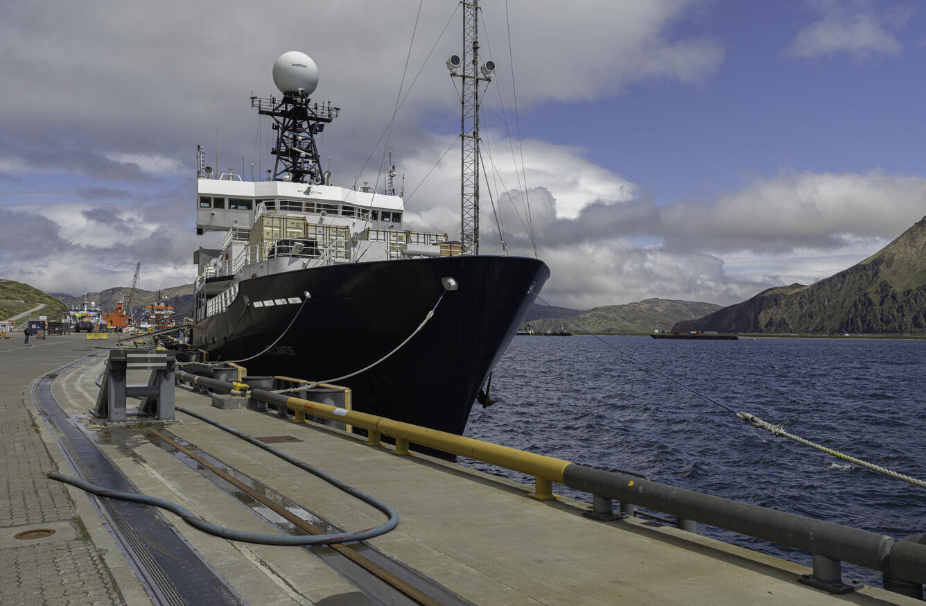 A research vessel at a dock with mountains, water, and blue sky with clouds in the background