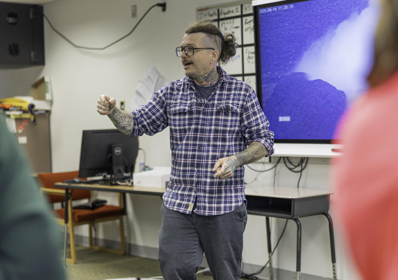 A person in glasses, plaid shirt, and jeans stands in front of a classroom