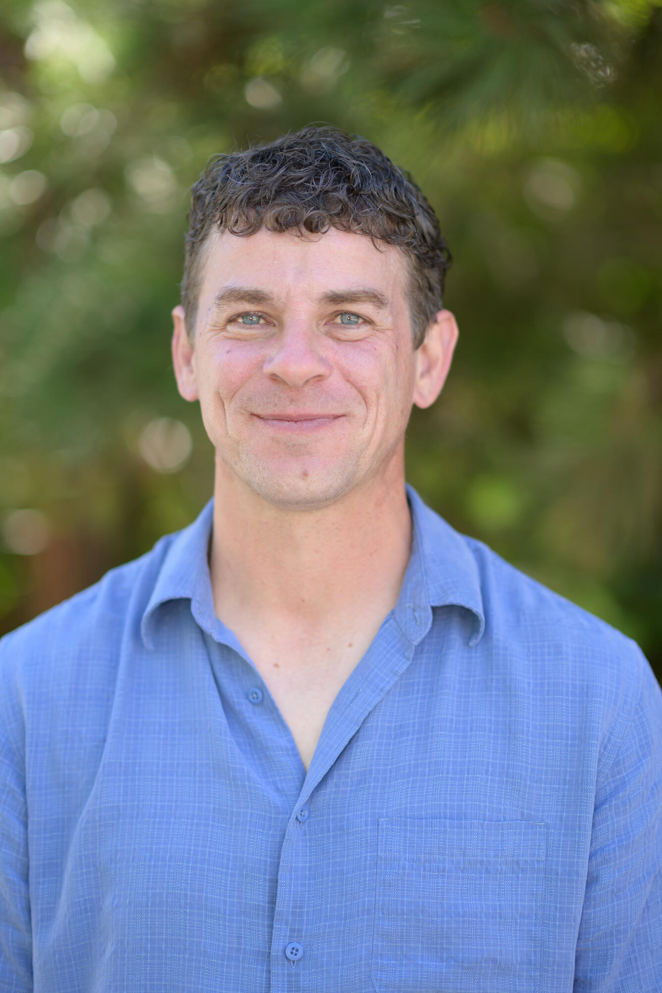 man with brown hair wearing a blue shirt standing near trees