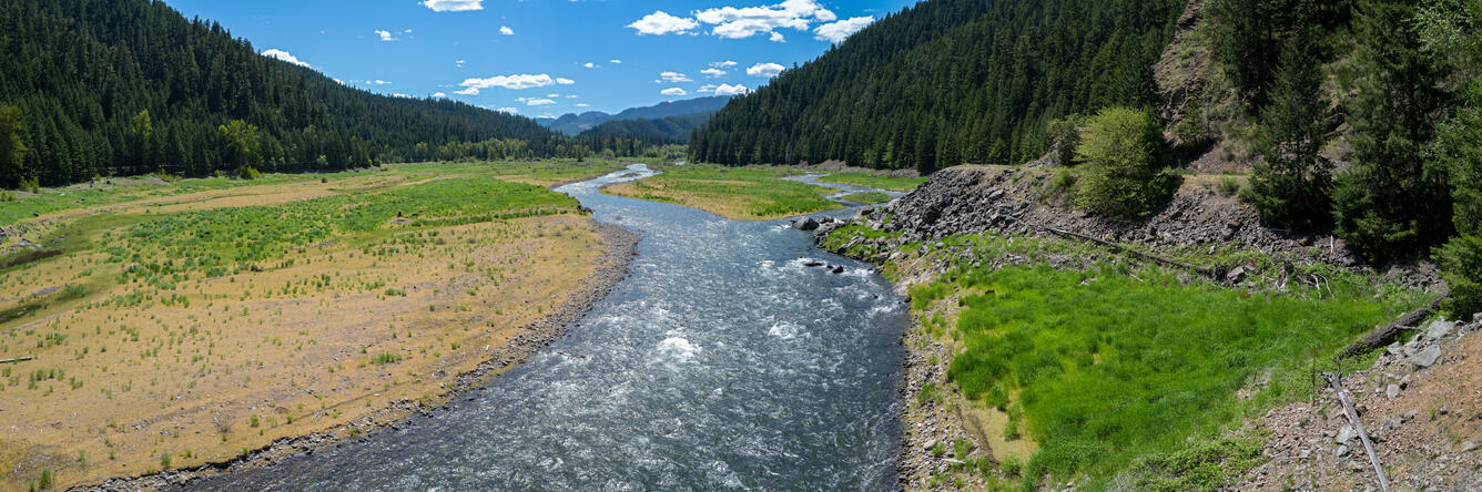 A blue strip of river runs through grassy, rocky fields. Steep hills with pine trees line both sides of the image