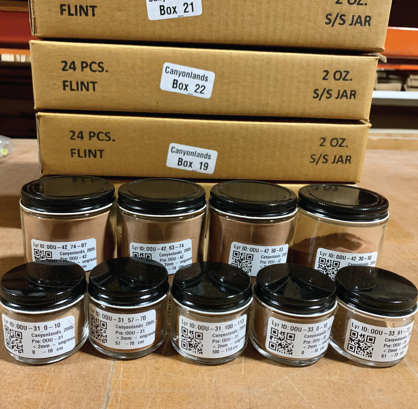 9 Jars full of collected soil samples in front of 4 stacked, cardboard boxes.