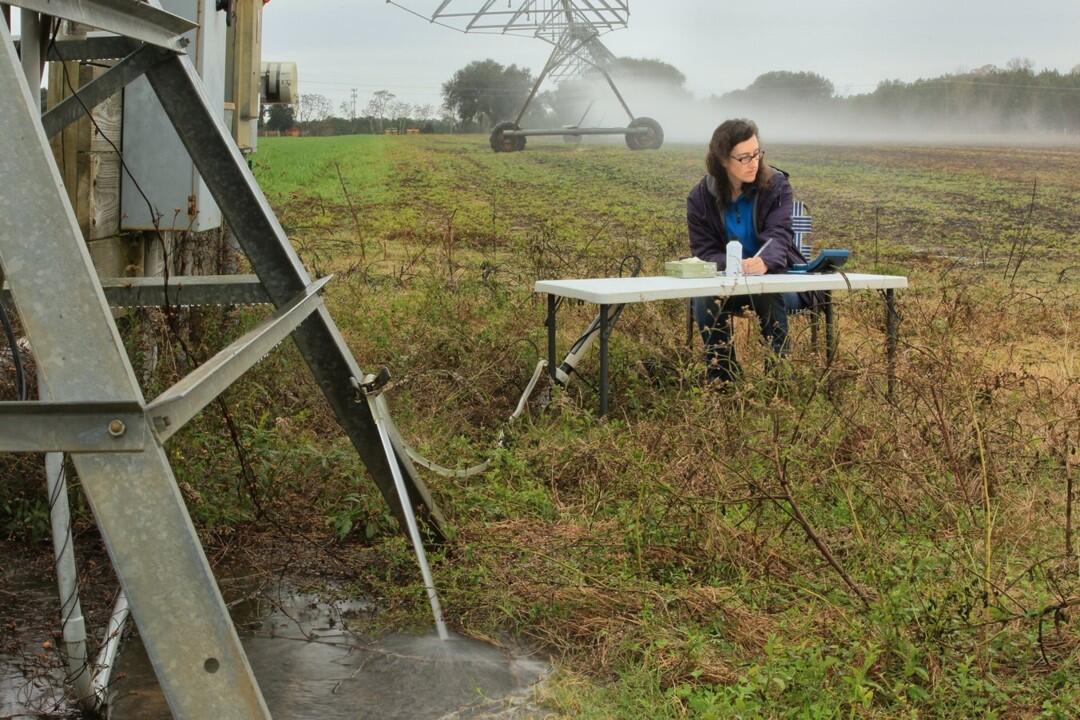 A scientist sits at a folding table in a field taking measurements from a well in the foreground