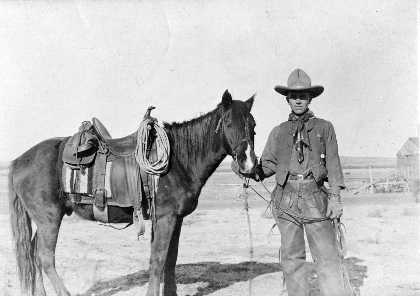 Black and white photo of man standing next to saddled horse