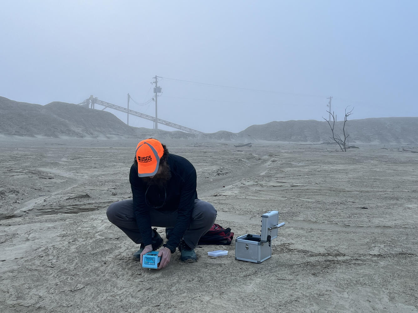 A man wearing an orange cap holds a device just above the ground to gather mine waste geophysical data