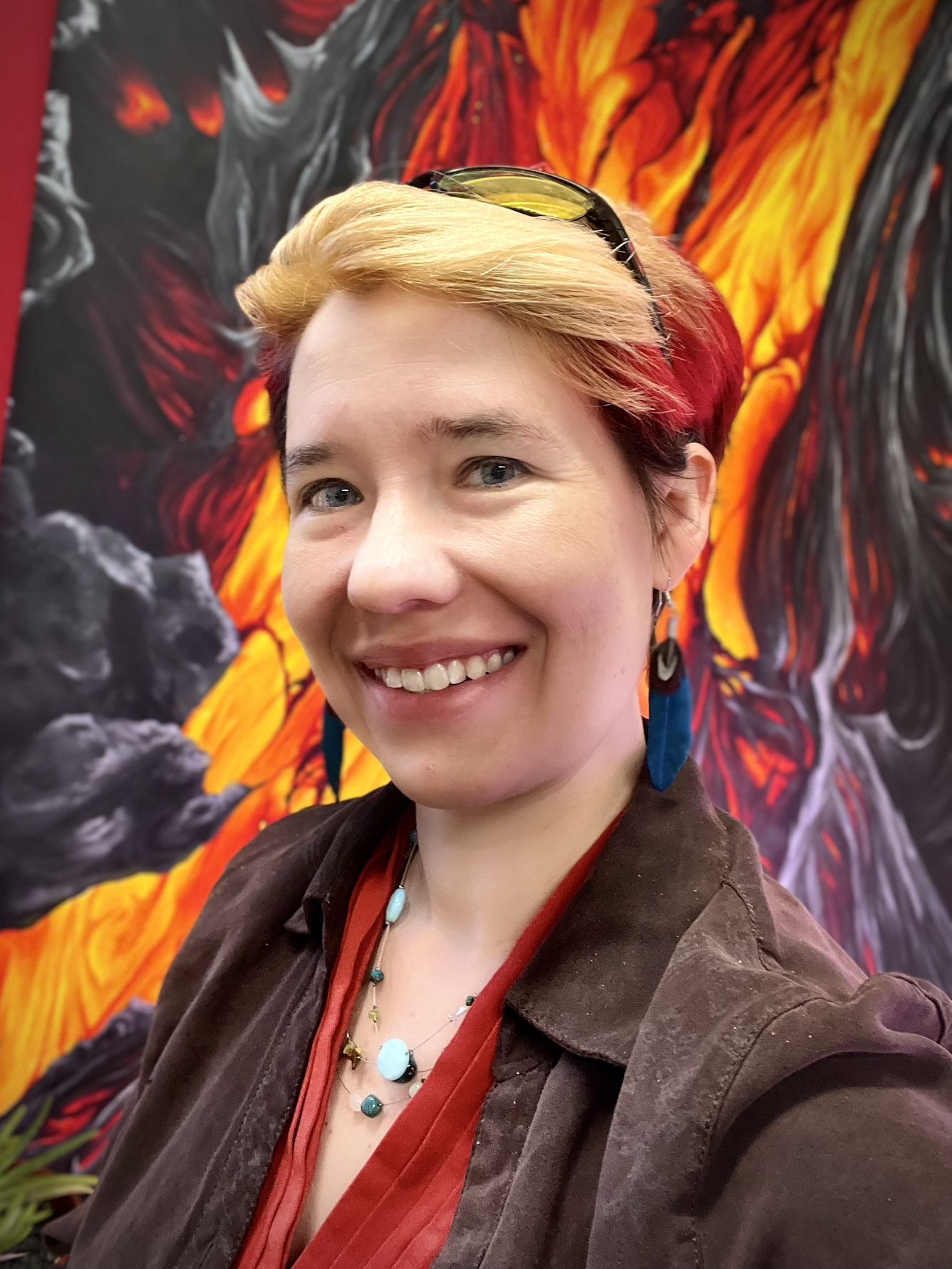 A woman with short blond-and-red hair, blue eyes, and a broad smile sits in front of a large painting of a glowing lava flow. She is wearing a bright red shirt and brown jacket.