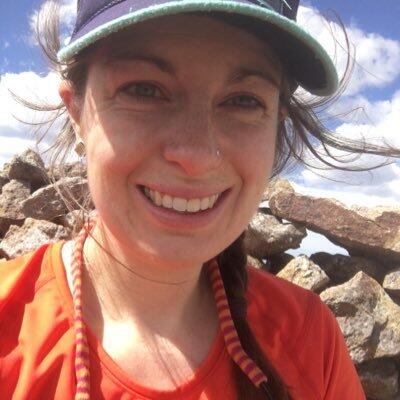 Alison Foster, smiling with rocks in the background. She is wearing an orange shirt and has a baseball cap on. 
