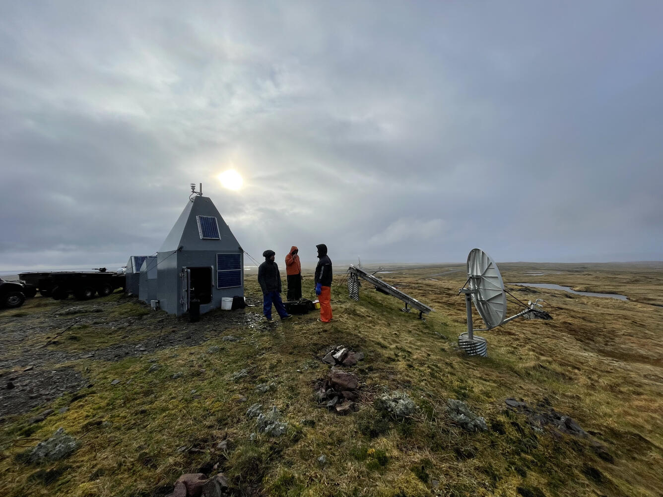 Three people stand beside a small hut on a flat plain, with a dish receiver in the foreground.