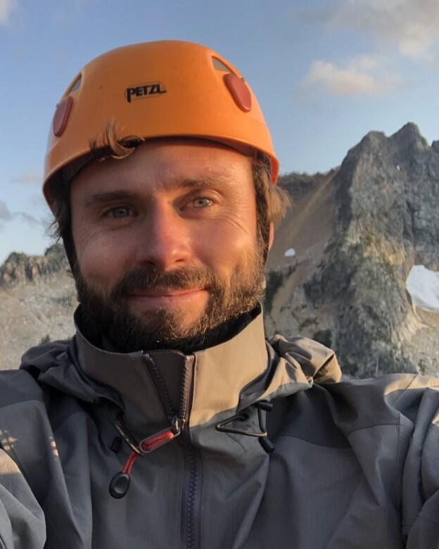 Headshot of smiling man wearing climbing helmet in front of mountain ridge