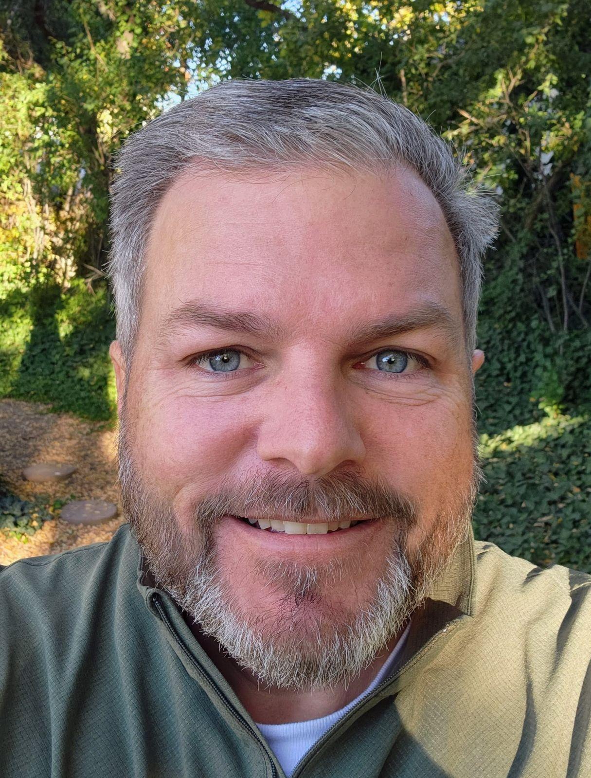 Photograph of Cory Angeroth wearing an olive green button up shirt with trees in the background.