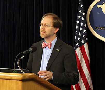 Image shows a man in a bowtie speaking at a podium with an American flag behind him