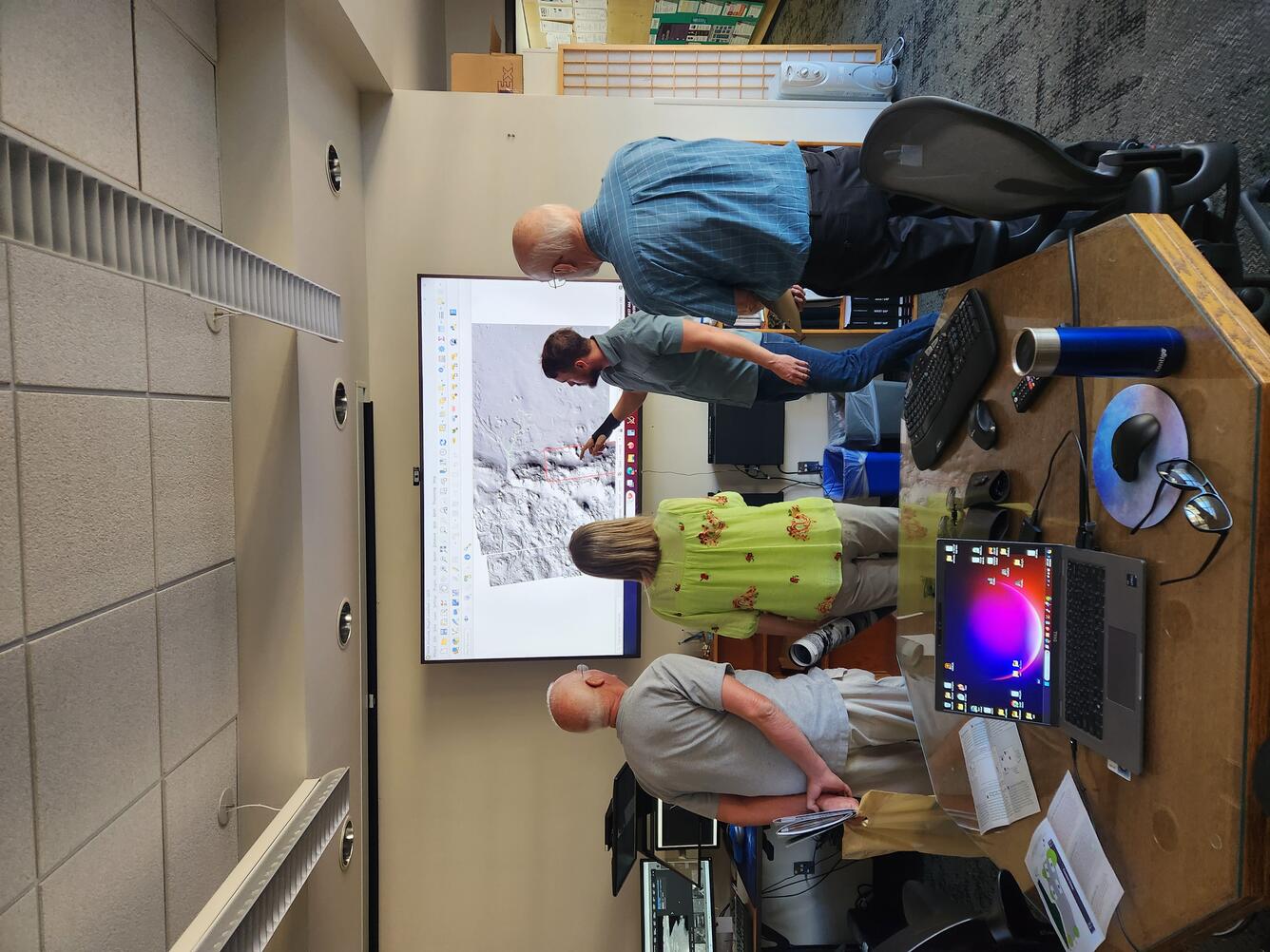 A man in a green shirt points to a planetary geologic map on a screen while engaging with the public at Astrogeology