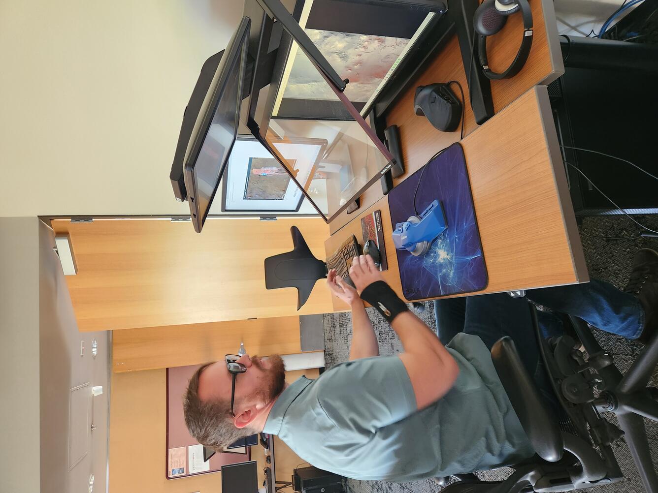 A man in a green shirt with glasses sits at a desk in the Astrogeology Photogrammetry Lab looking at monitor screens.