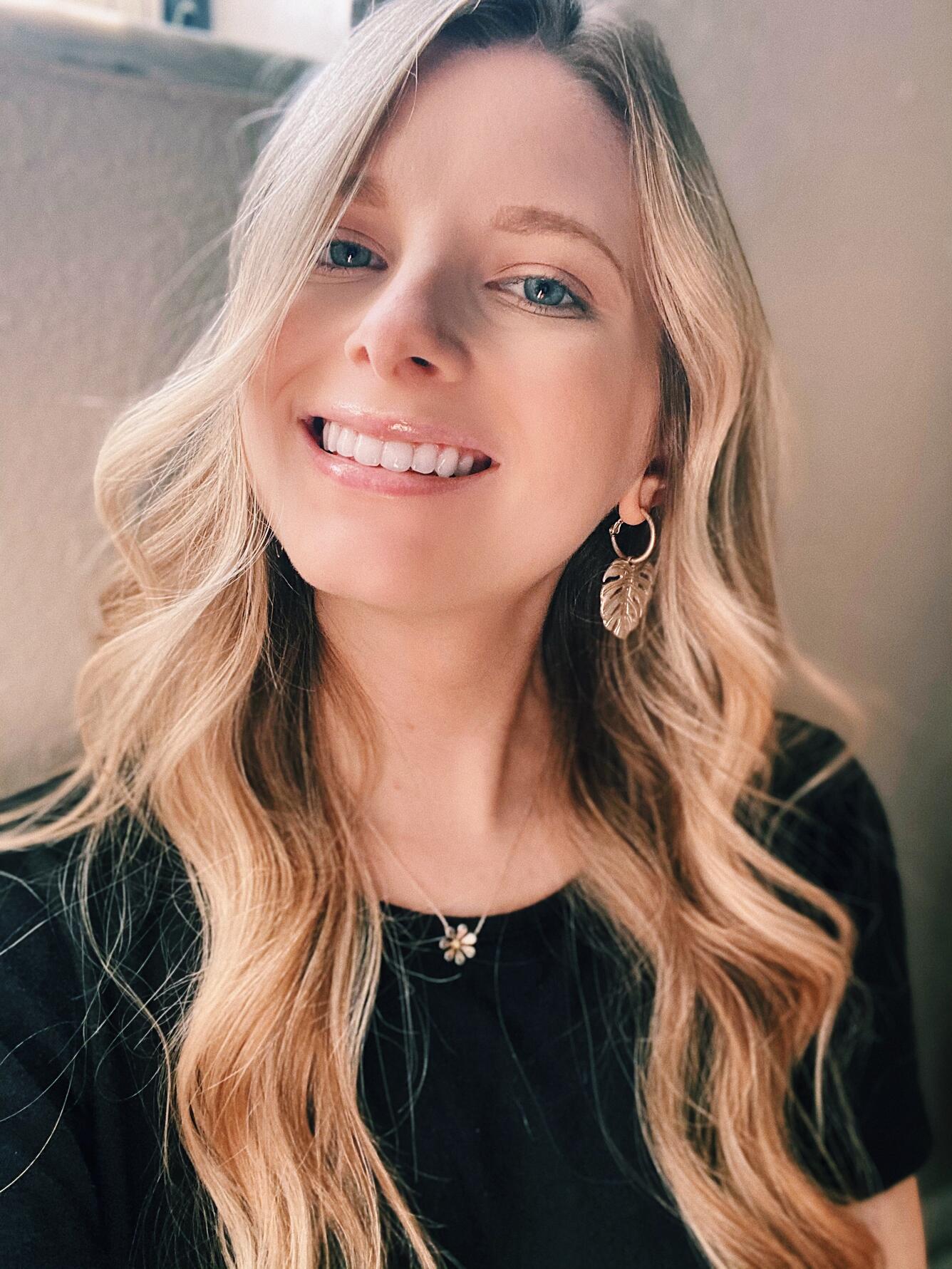 A woman with blond hair and a black blouse stands against a tan background.