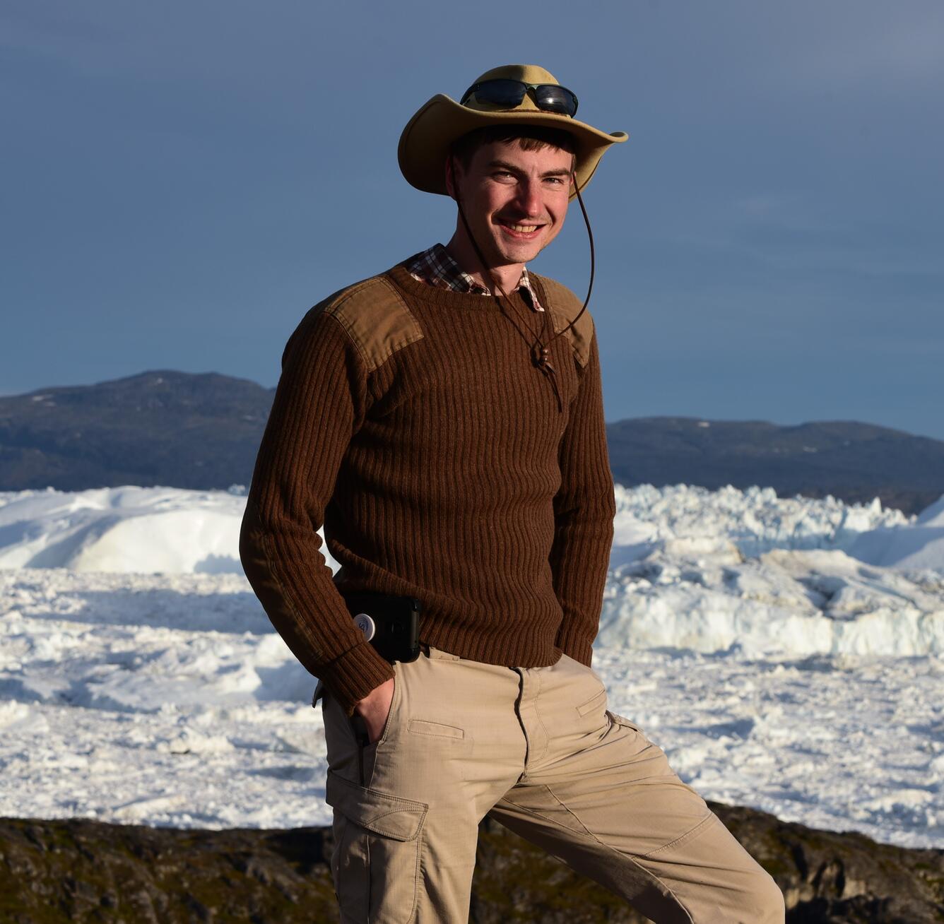 Photo of Brandon Lars Graham wearing winter cloths, hat, and sunglasses. Snow and mountains are in the background.
