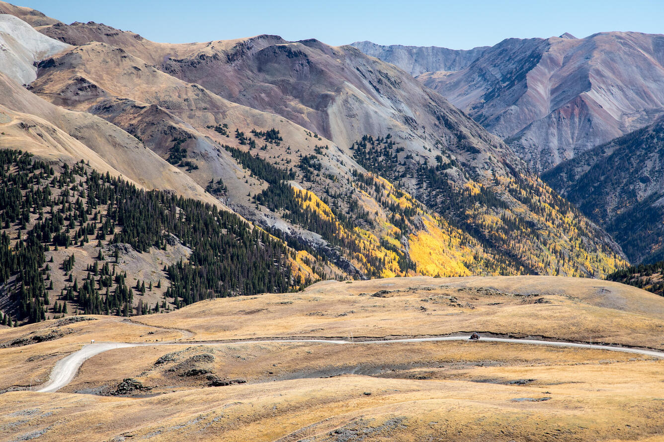 Mountains with brown grasses, yellowing trees with a field and road in front 