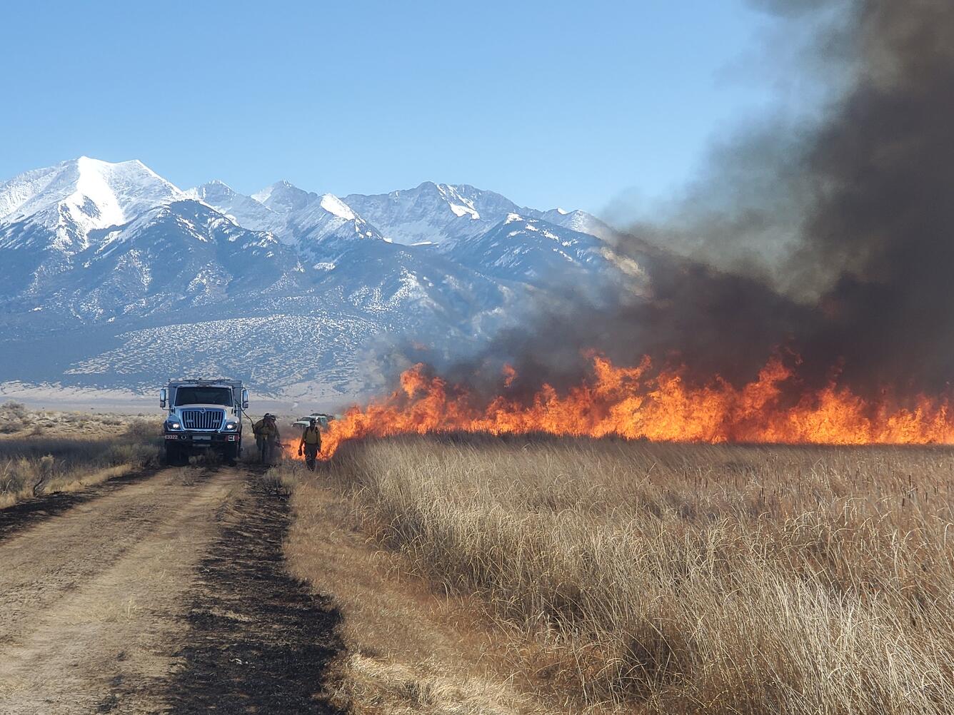 People standing near a fire, white truck, and snow-capped mountains in the background 