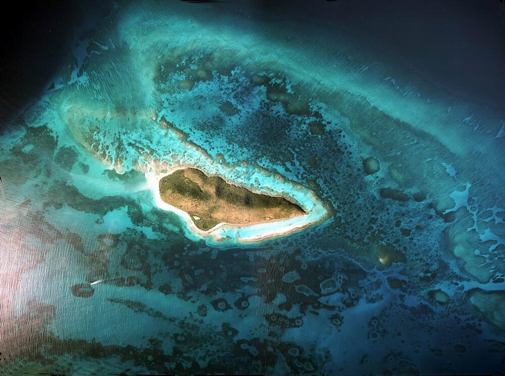 view from space looking down at a teardrop shaped green island lined with white beaches, turquoise water, and coral reefs