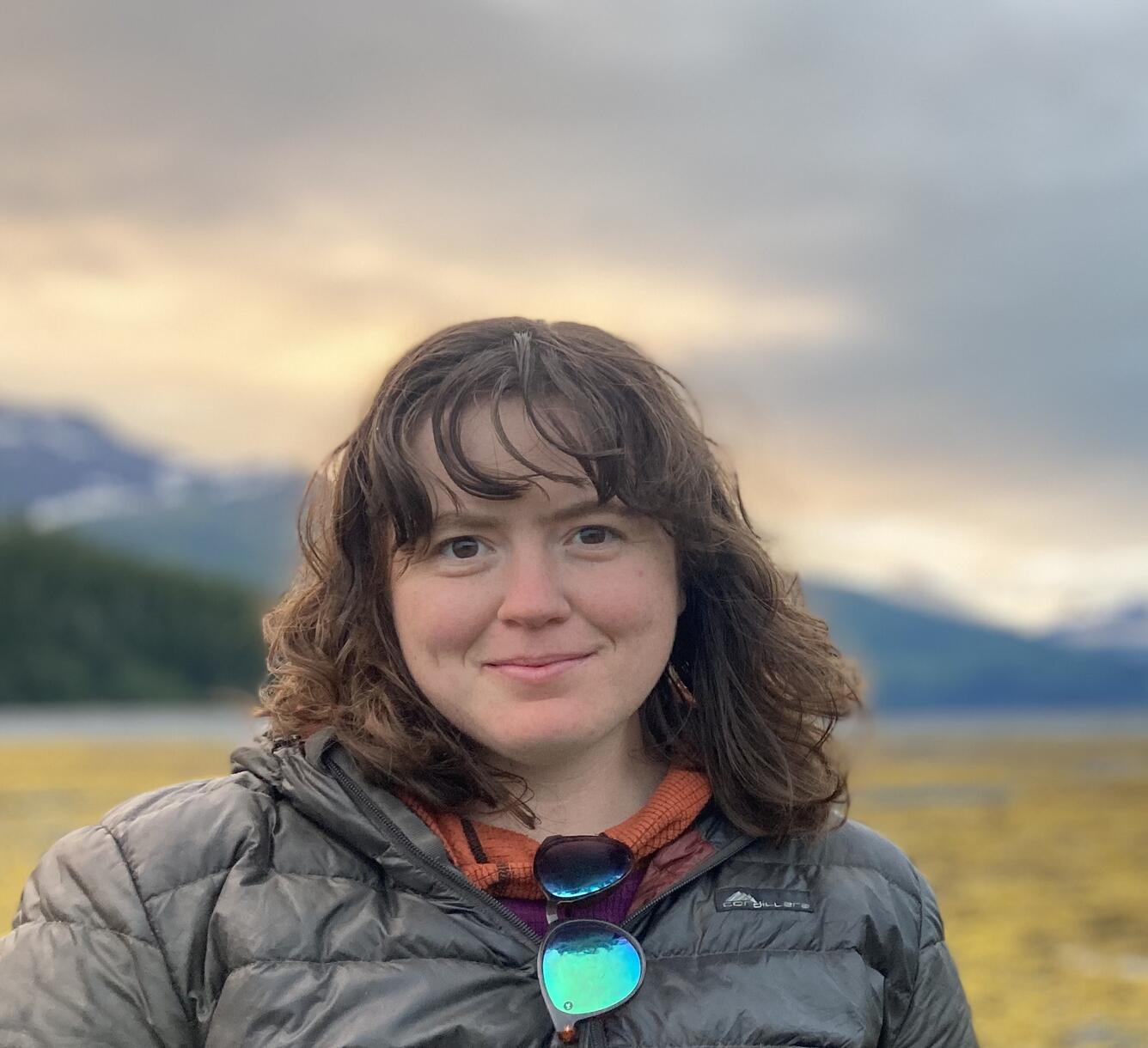 A woman with brain hair and black puffer jacket stands in front of a blurry mountain scene