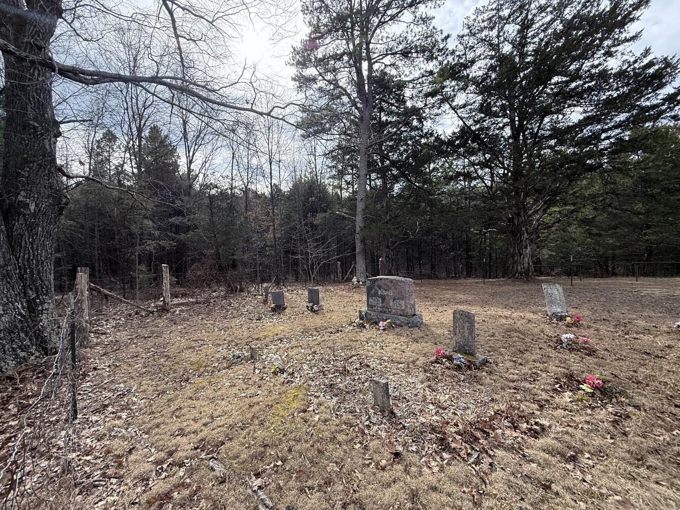 A small, quiet cemetery sits in a clearing bordered by forest, with several old headstones and flower arrangements