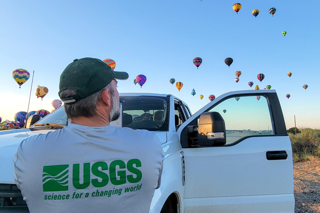 A USGS employee standing in front of their work vehicle looks at the dozens of hot air balloons rising in the sky