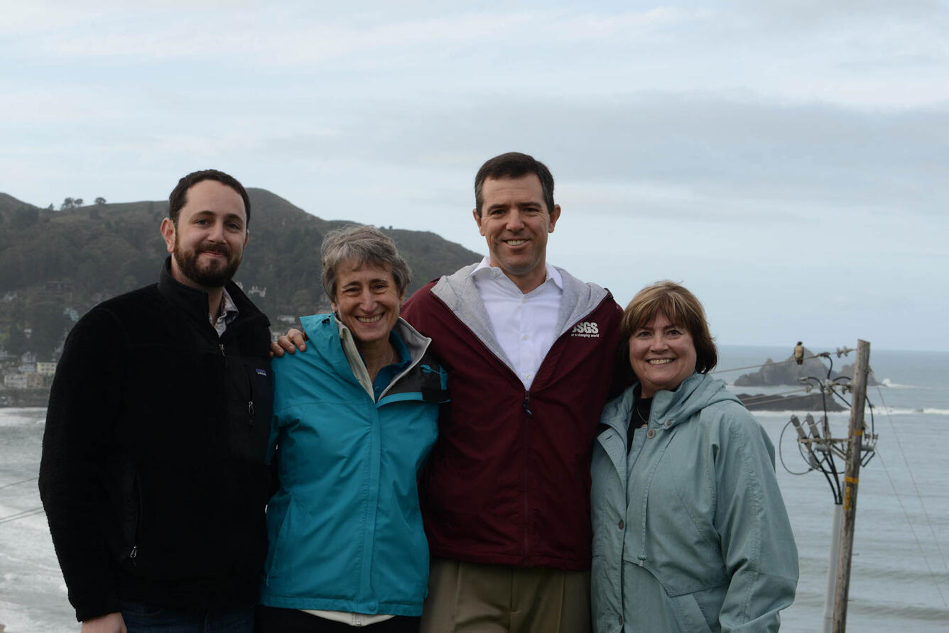 Four people, 2 men and 2 women, stand together arm-in-arm, smiling for the camera.