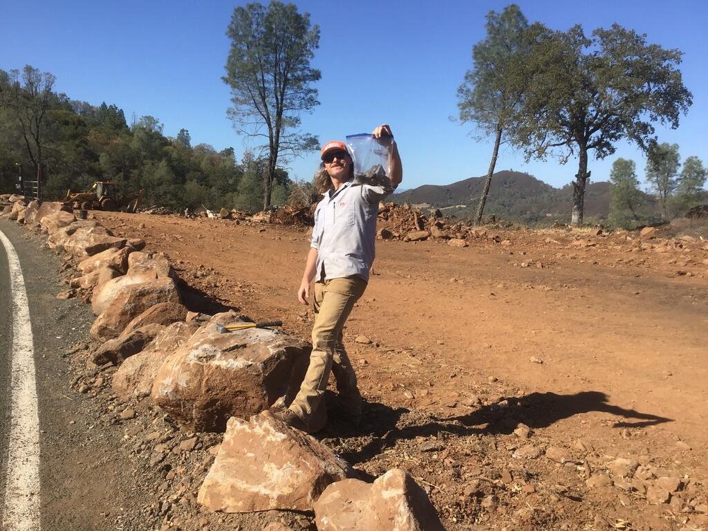 A geologist in tan pants, a blue button-down shirt and an orange hat stands with one foot on a boulder at the side of a road, holding up a baggie of rocks. Behind him the ground has been scraped bare by construction equipment.  