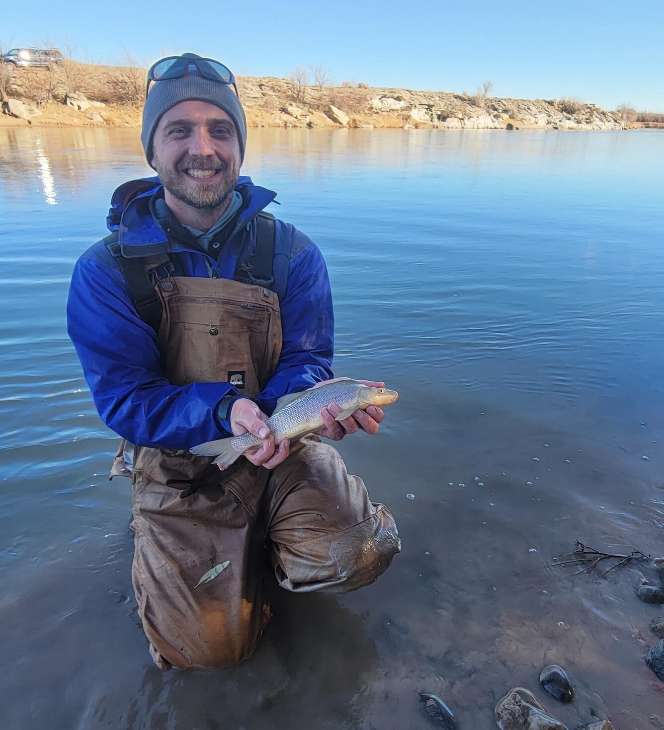 graduate student standing near blue water, wearing a black hat, black sunglasses and a blue sweatshirt, holds a razorback sucker fish 