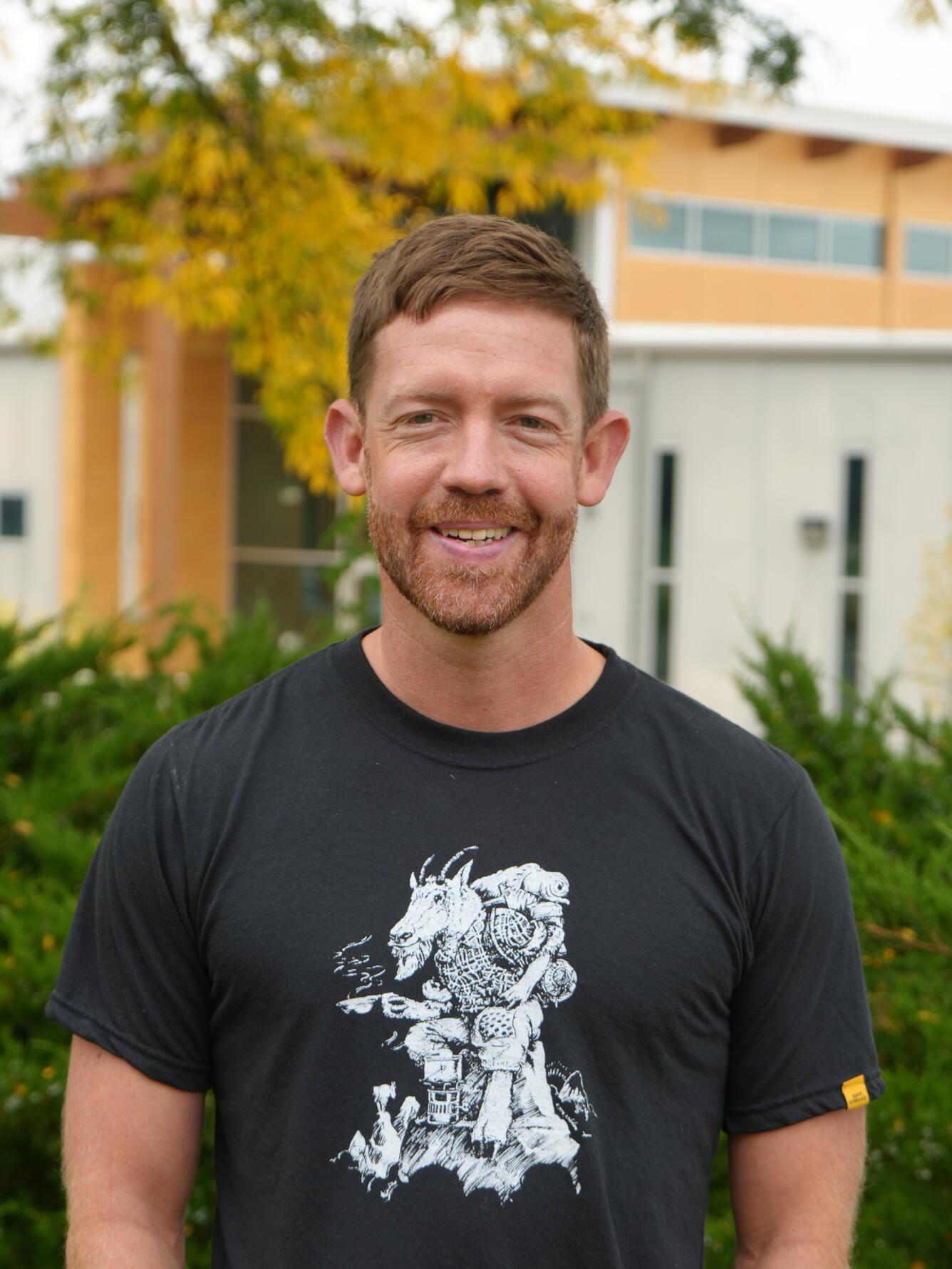Man with brown hair and short beard smiling with yellow-leafed tree and modern building in background
