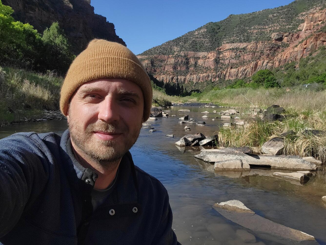 A man wearing a beige cap looks directly at the camera with a stream and red rock canyon behind him