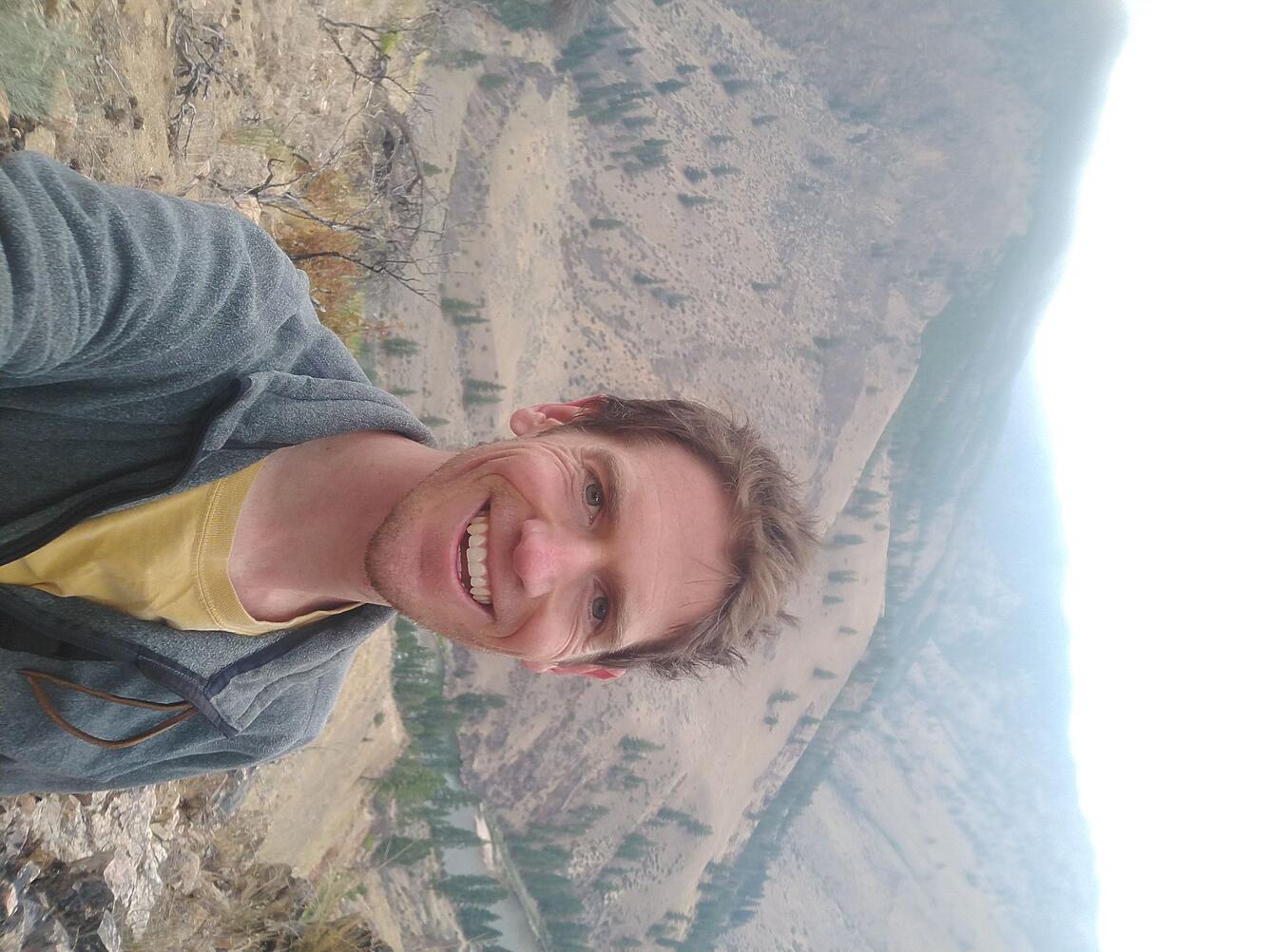 Photo of Benjamin Robb, smiling with mountains in the background. Benjamin is wearing a yellow t-shirt and gray sweatshirt.  