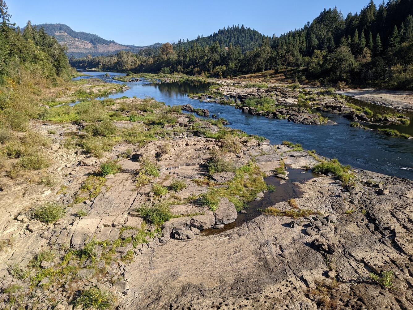 a wide, rocky riverbed with shallow, clear water in the foreground. Rolling forested hills and blue sky in background