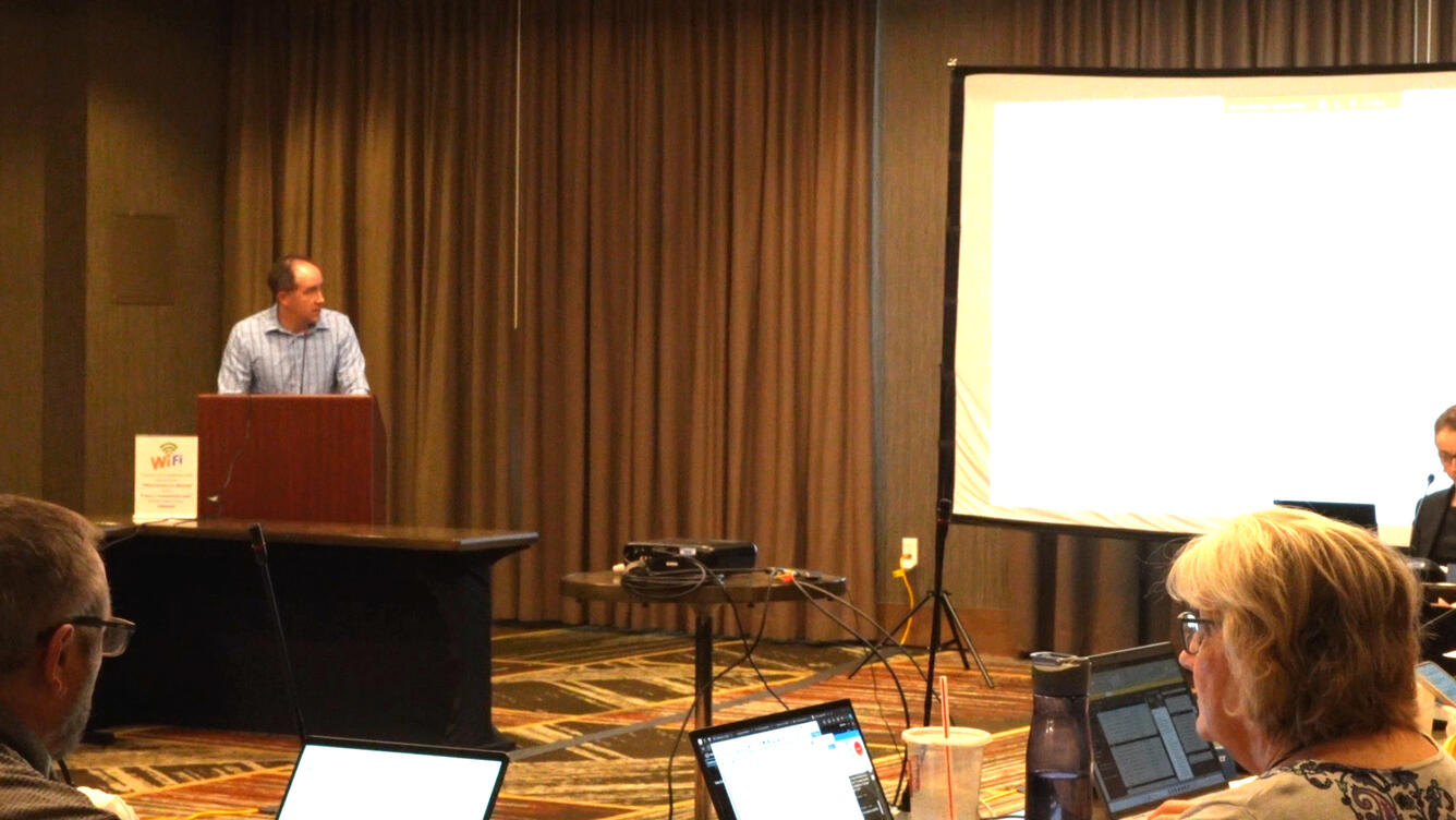 A balding man presents a PowerPoint from a lectern in front of tables of people bent over laptops.