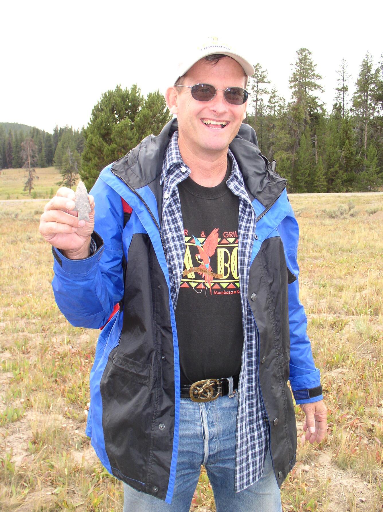 Photo of a man in sunglasses, a rainjacket, and jeans, holding up an arrowhead