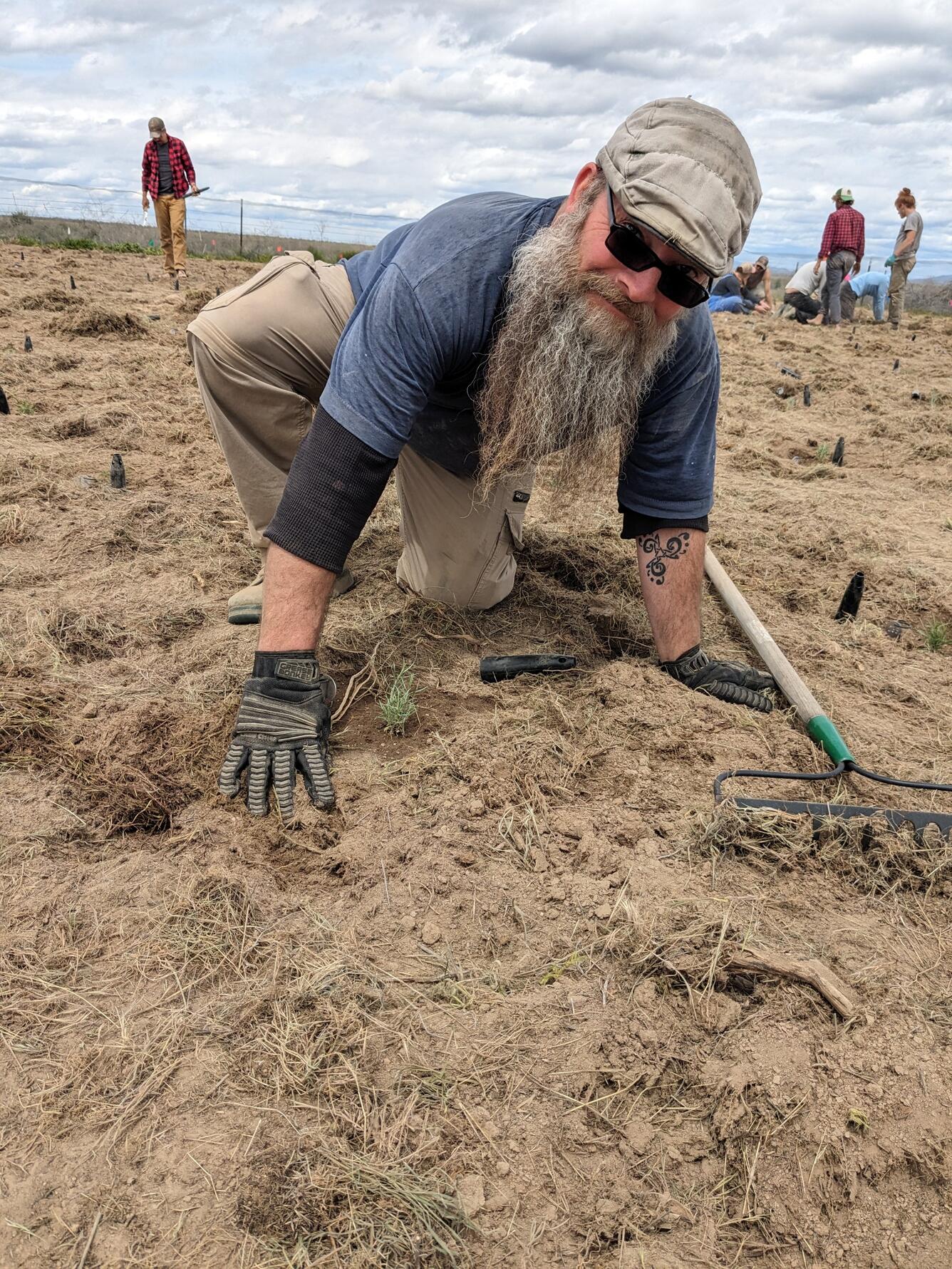 Bill touching the dirt with a rake at his side. Hes wearing gloves and has a long beard, sunglass, and a hat on
