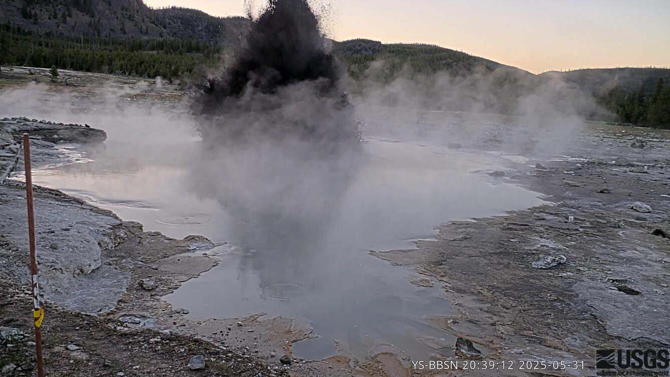At sunset a small plume of muddy water erupts from an otherwise placid, steaming pool surrounded by barren rocky ground